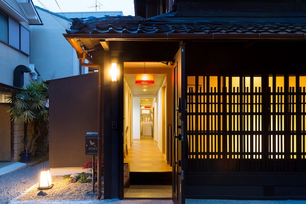 A traditional Japanese gate with a wooden lattice design, a post office sign, and a pathway leading to a building.