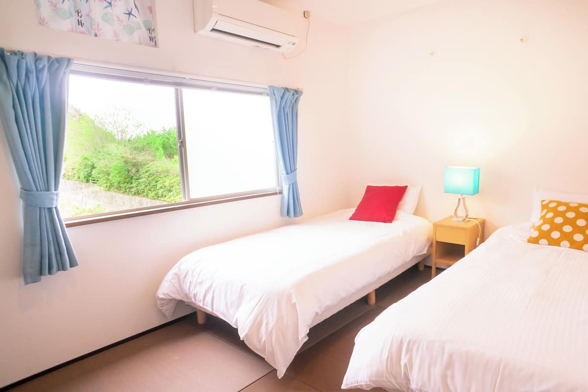 A room with two beds, a window with curtains, a bedside table with a lamp, and an air conditioning unit.