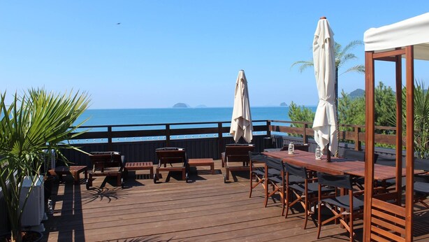 A wooden deck with outdoor seating, umbrellas, and a view of the sea.