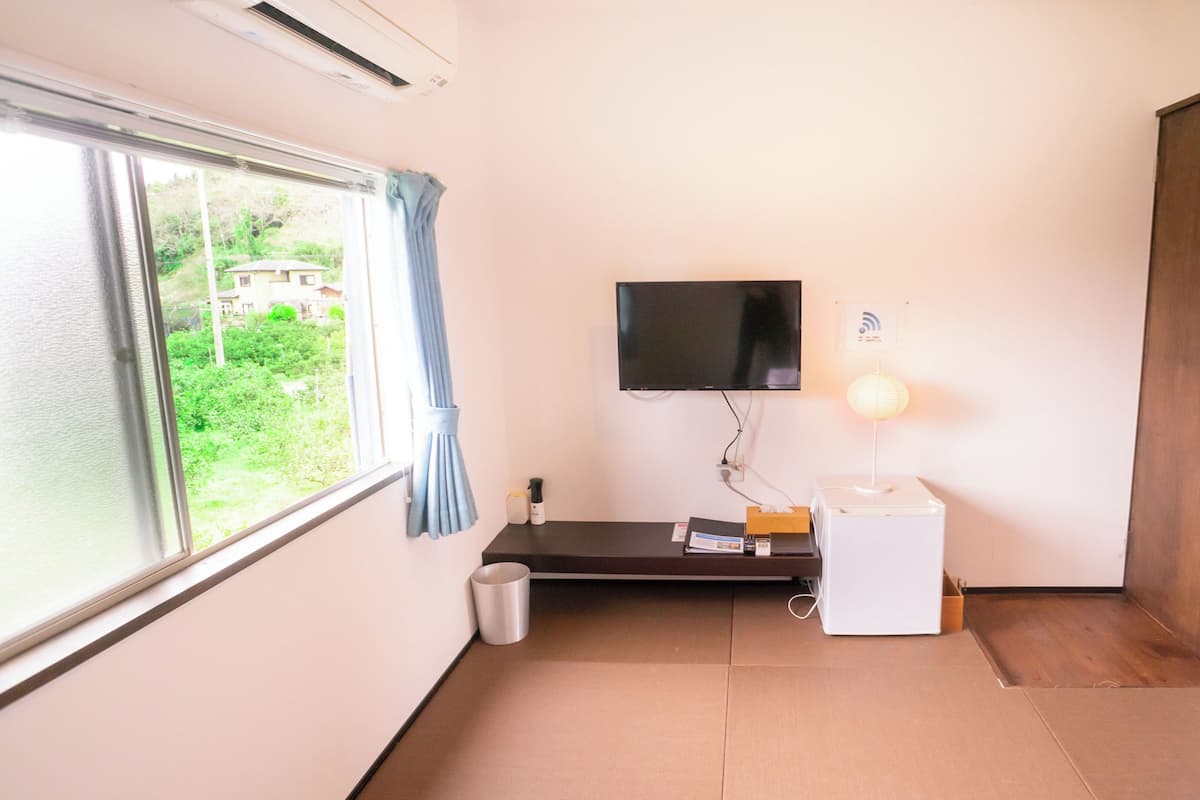 A room with a TV, a bedside table, a lamp, a waste bin, and a window with curtains.