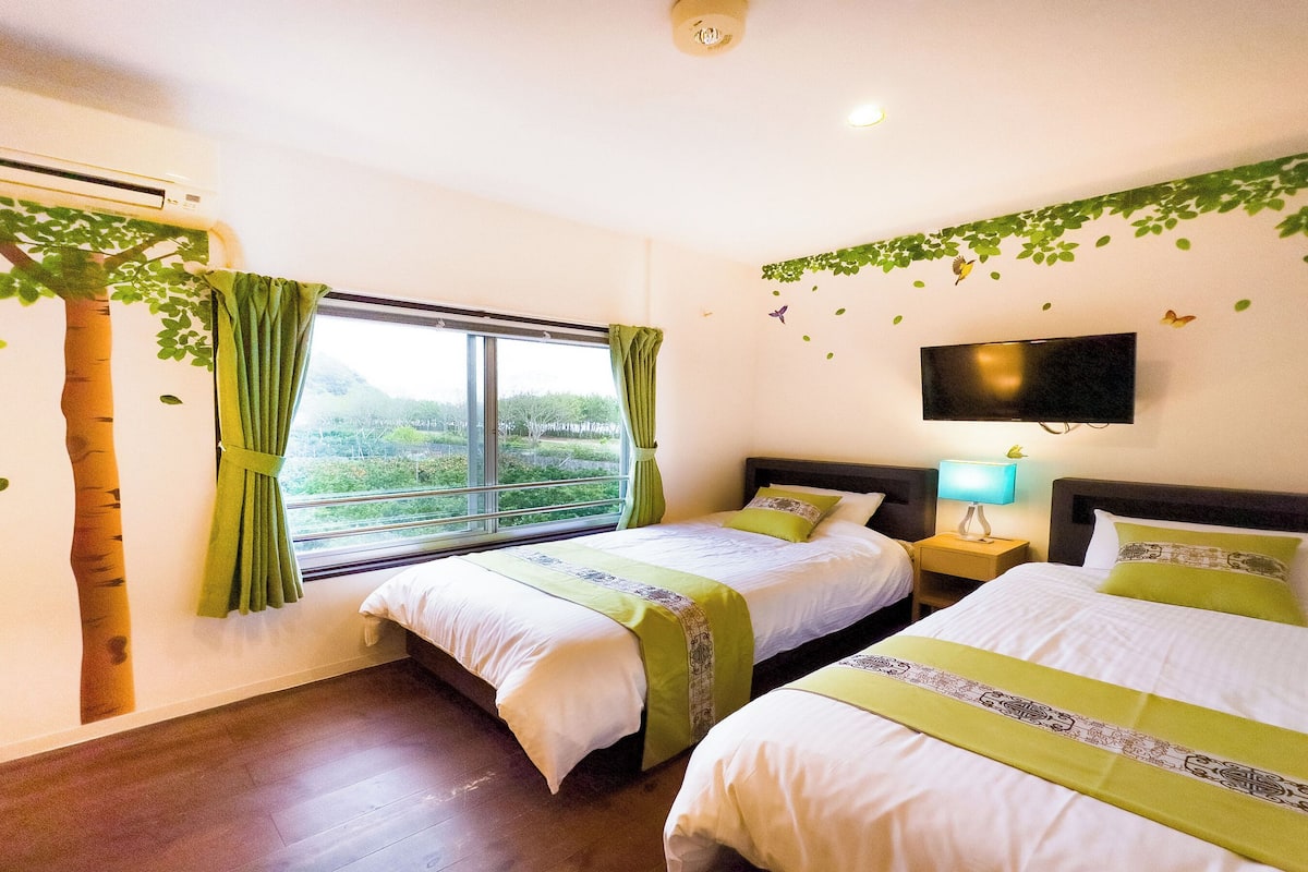 A hotel room with two beds, a TV, a window with curtains, and a view of greenery outside.