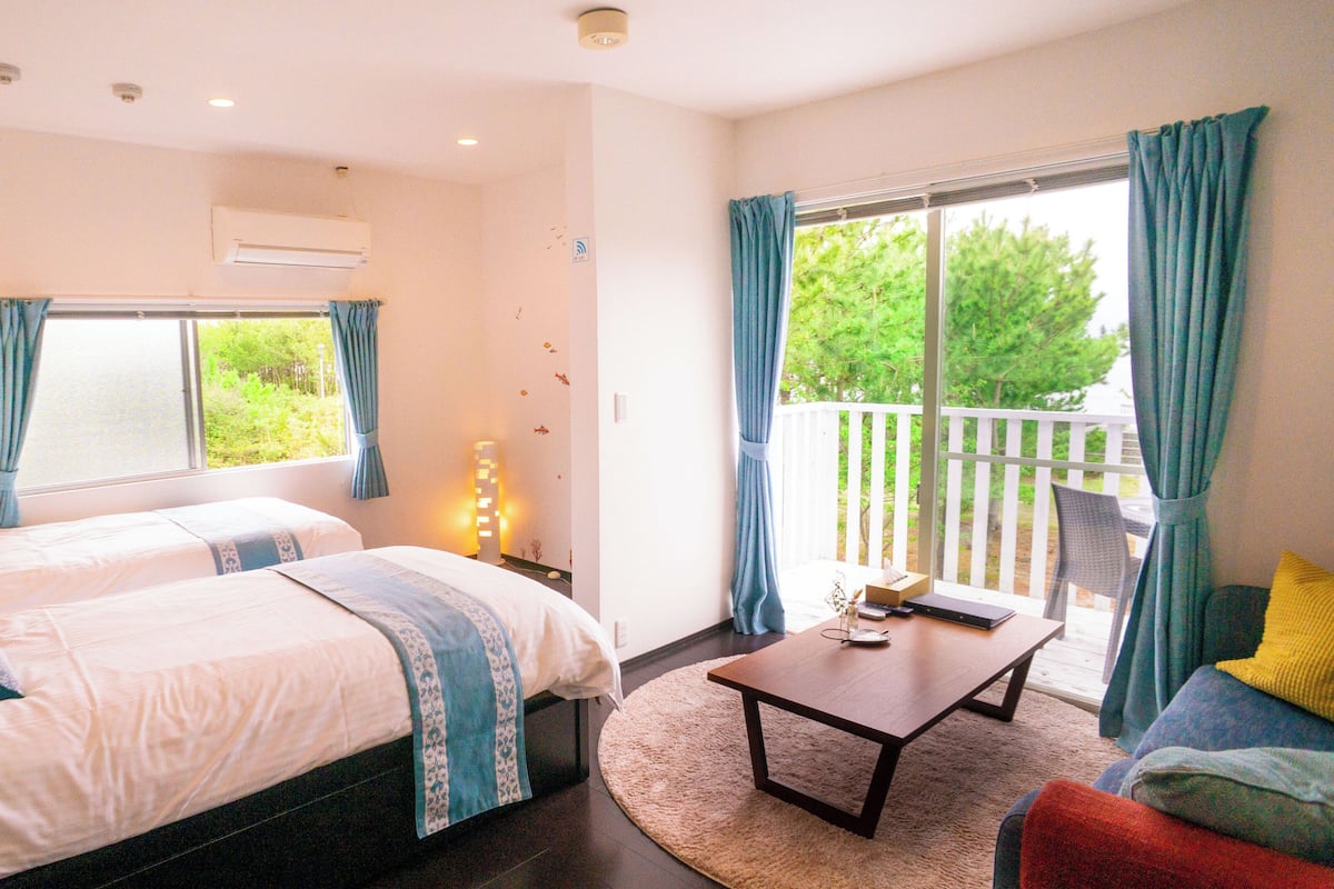 A bedroom with two beds, a bedside table with a lamp, a small table with a book, and a view of the outdoors through a sliding door.