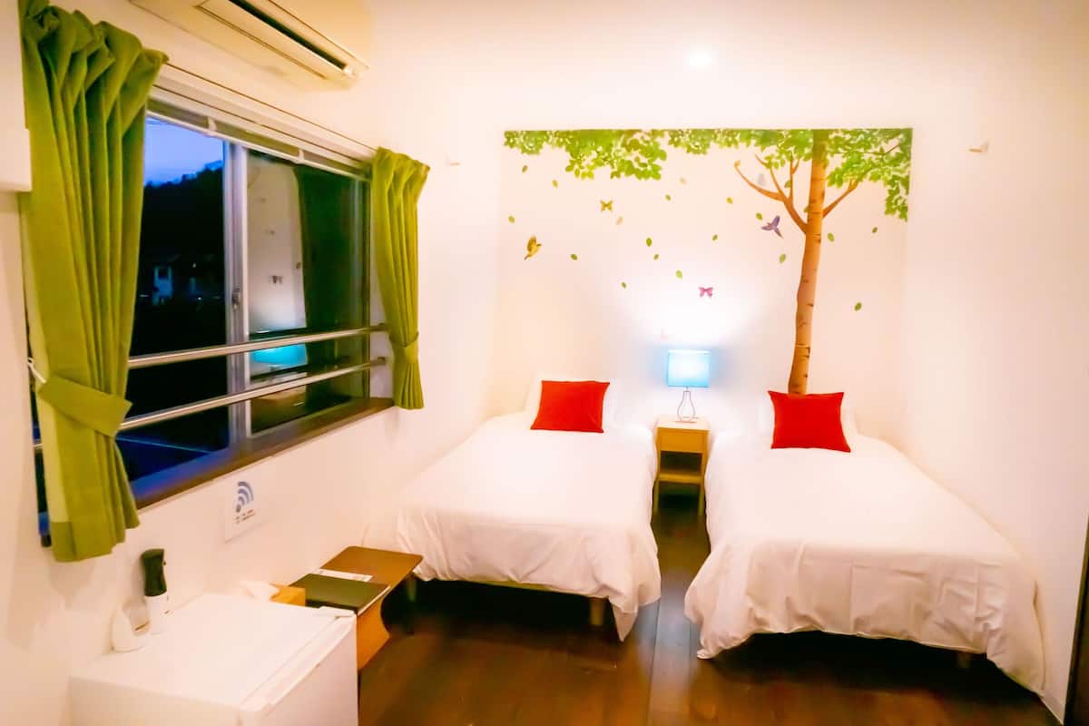 A hotel room with two single beds, a window with curtains, a wall-mounted air conditioner, a small table with a lamp, and a wall decoration of a tree.