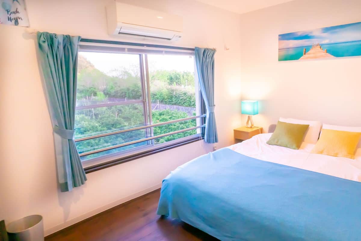 A bedroom with a bed, a nightstand, a lamp, a painting, and a window with curtains.