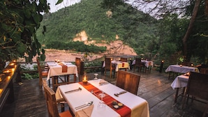 An outdoor dining area with tables set for two, surrounded by wooden chairs and tables, overlooking a forested hillside. - Muang La Lodge (Muang La)