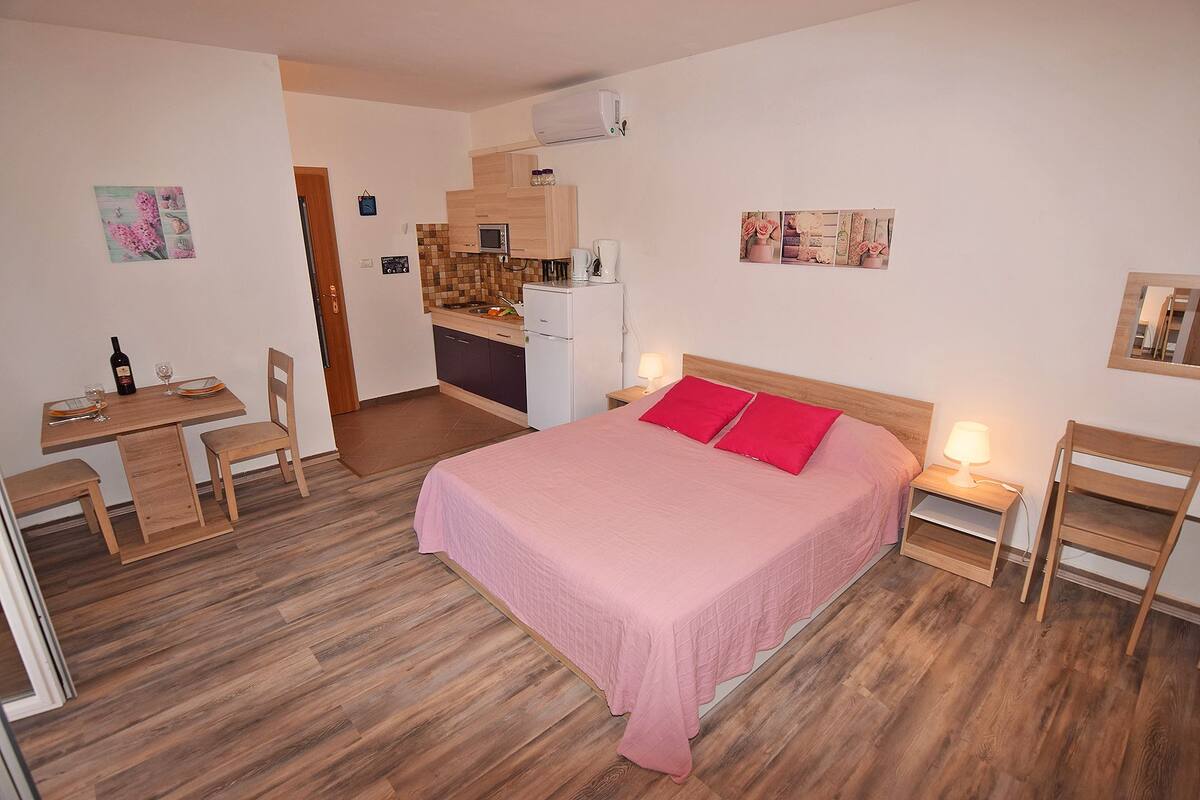 A compact bedroom with a bed, bedside tables, a desk, and a kitchenette.