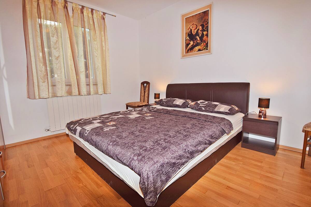 A bedroom with a large bed, a nightstand, a chair, a painting on the wall, and a window with curtains.