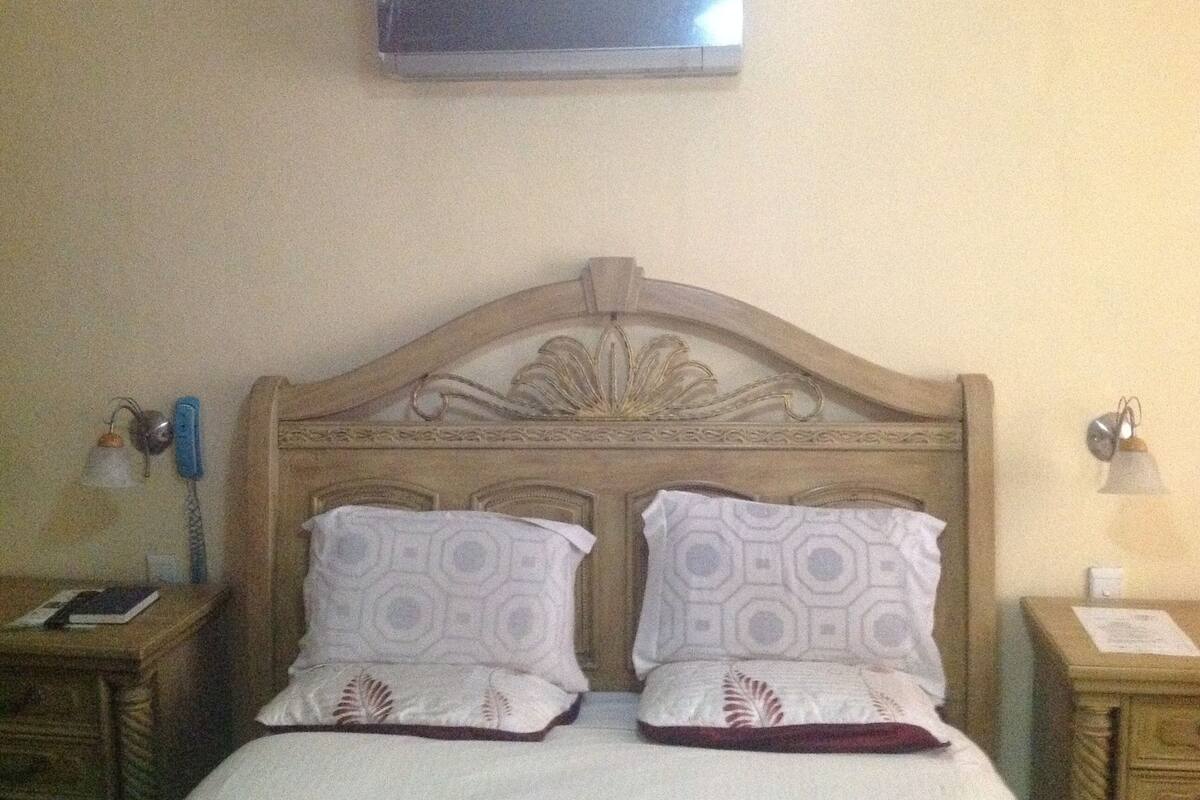 A hotel room with a wooden headboard, two patterned pillows, a flat-screen TV mounted on the wall, and bedside lamps.