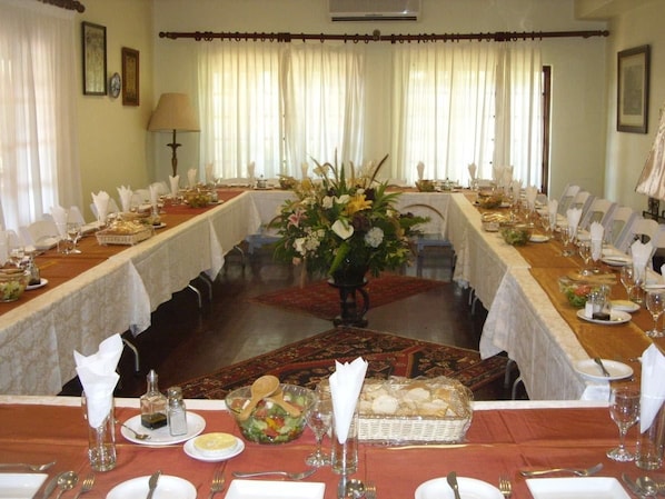 A formal dining room with a long table set for a meal, surrounded by chairs, with a floral centerpiece and a patterned rug.