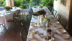 A long dining table set for a meal with chairs on both sides, surrounded by greenery and a brick floor.