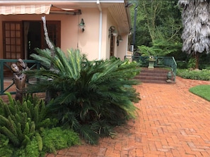 A brick pathway leading to a building with a porch, surrounded by lush greenery and a thatched roof structure.