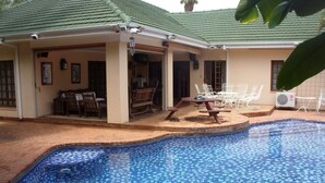 A swimming pool with a tiled pattern, a covered patio area with outdoor furniture, and an air conditioning unit.