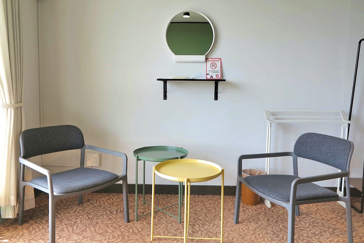 Una habitación con dos sillas grises, una pequeña mesa redonda con base amarilla, una mesa verde y un espejo en la pared.