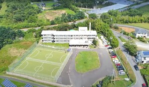 An aerial view of a school with a large sports field, a parking area, and surrounding greenery.