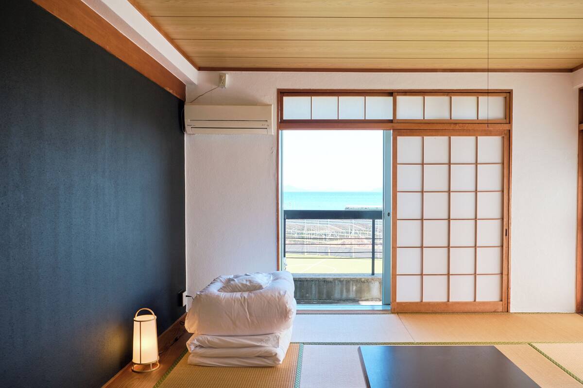 Una habitación con suelo de tatami, una cama futón, un farol y una puerta corredera que da a un balcón con vistas al mar.