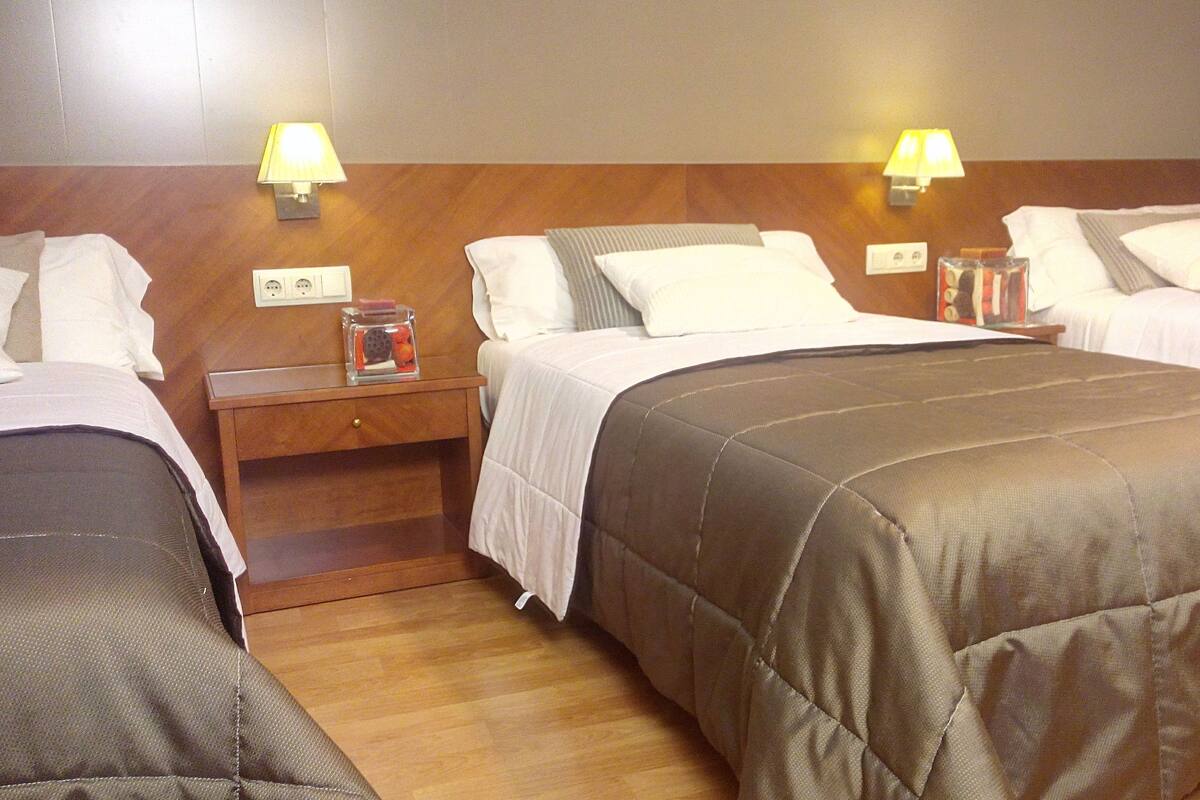 A hotel room with three single beds, each with a wooden headboard, white bedding, and a nightstand with a lamp.