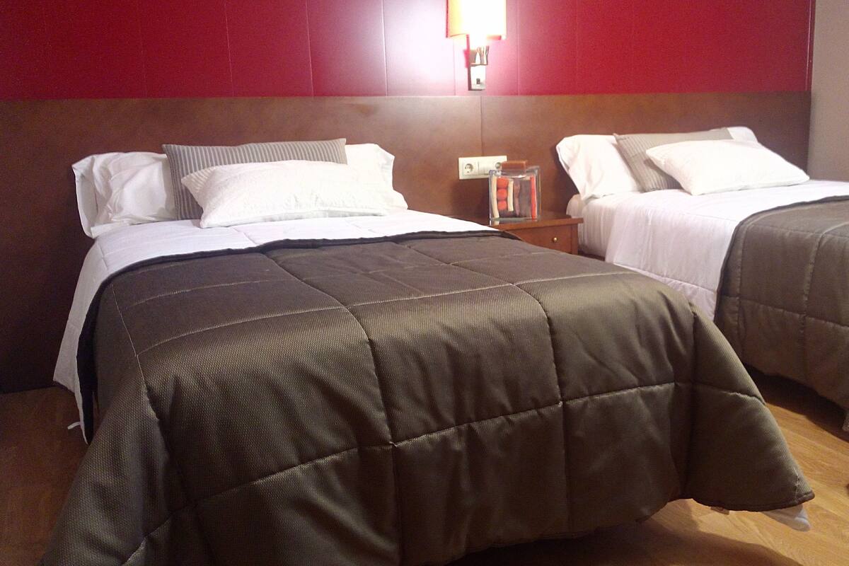 A hotel room with two beds, each with a dark-colored comforter and white pillows. There is a nightstand with a lamp and a small table with books and a decorative item.