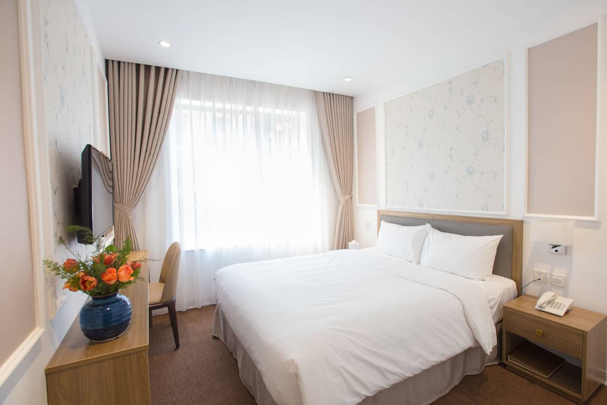 A hotel room with a large bed, a bedside table with a lamp, a vase with flowers, a chair, and a TV on the wall.