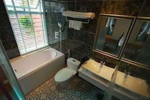 Une salle de bain équipée d’une baignoire, de toilettes et d’un lavabo, avec des carreaux de sol à motifs et une fenêtre avec des stores.