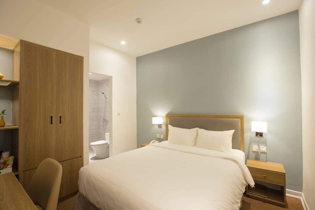 A hotel room with a bed, a wooden wardrobe, a desk, and a nightstand with a lamp.