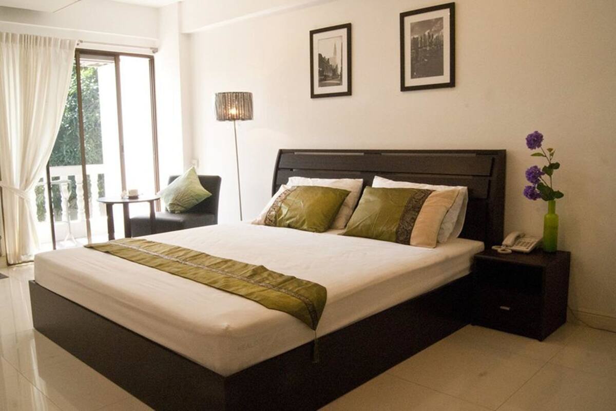 A hotel room with a large bed, a bedside table with a lamp, and a view of the outdoors.