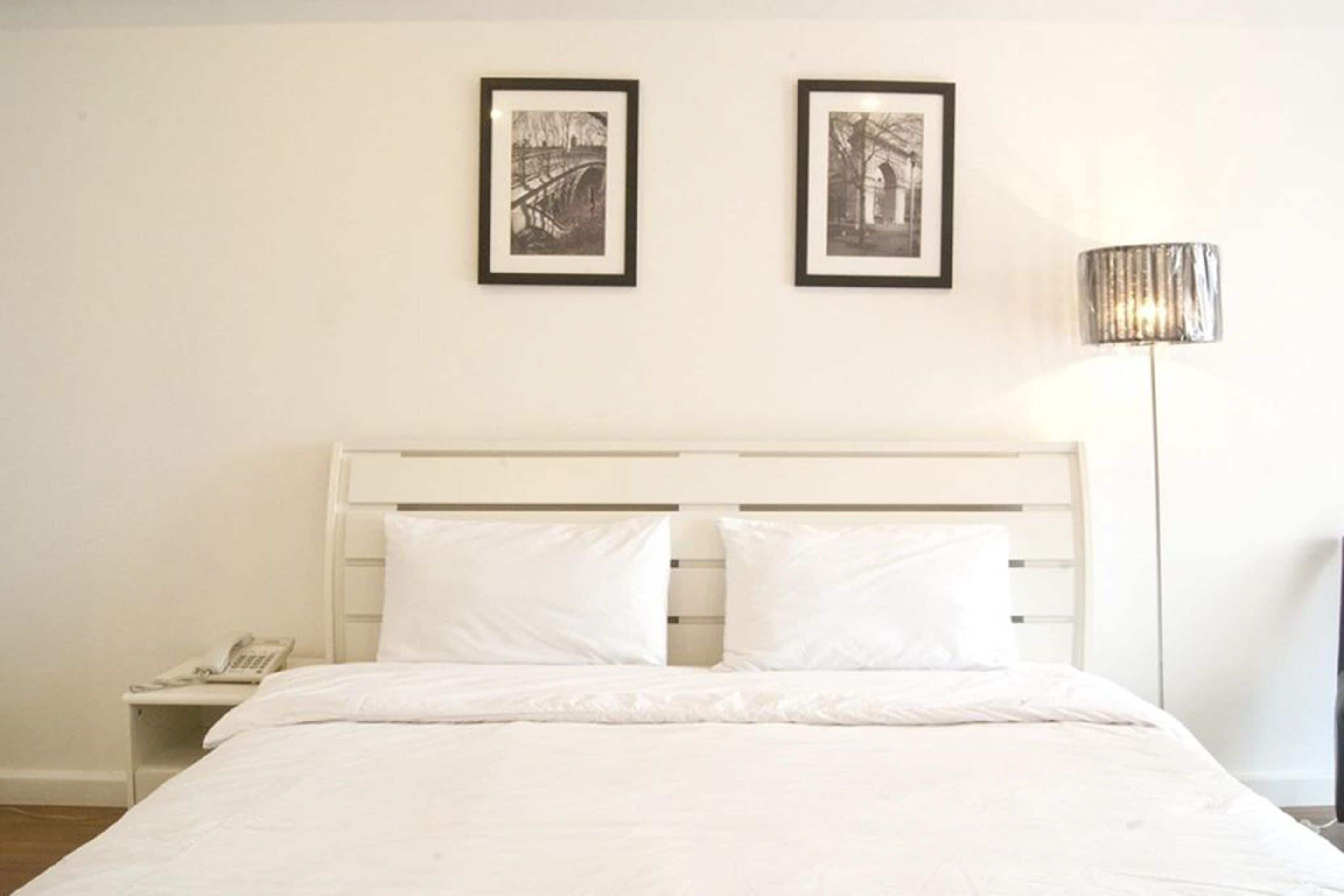 A neatly made bed with white bedding, a bedside table with a phone, and two framed pictures on the wall.