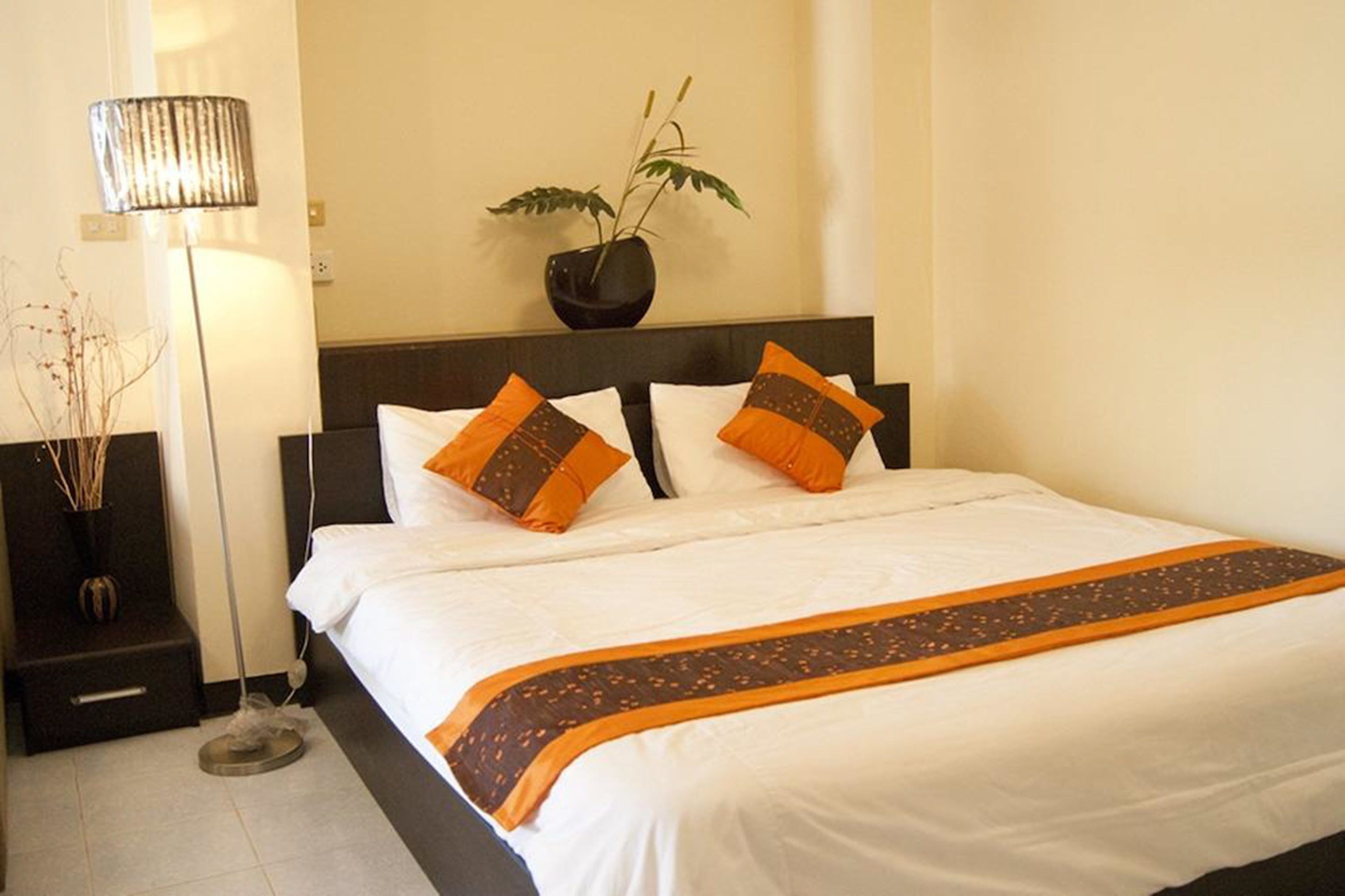 Una cama bien hecha con ropa de cama blanca y naranja, un cabecero oscuro y una planta en maceta en la mesita de noche.