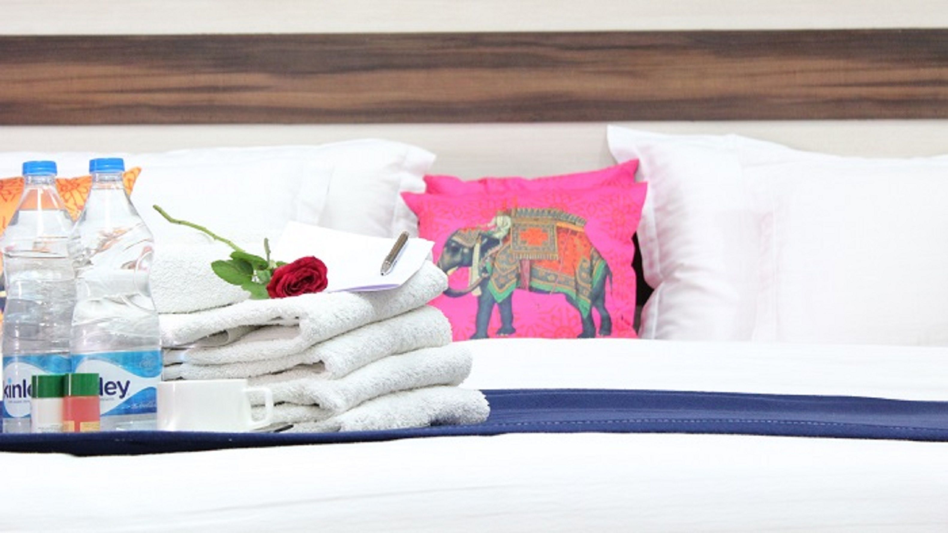 Una cama bien hecha con sábanas blancas, un almohadón rosa con diseño de elefante y una falda de cama azul. Hay dos botellas de agua y una rosa sobre la cama.
