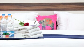 Una cama bien hecha con sábanas blancas, un almohadón rosa con diseño de elefante y una falda de cama azul. Hay dos botellas de agua y una rosa sobre la cama.