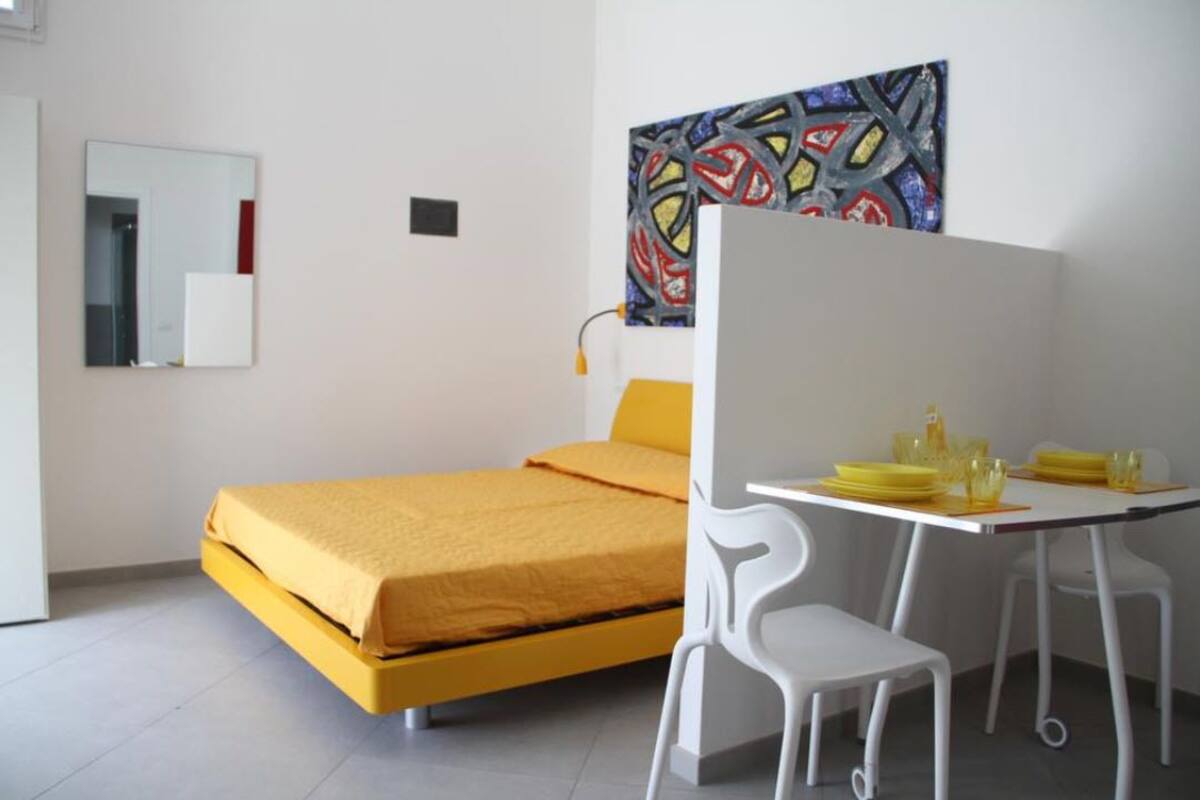 Un dormitorio con una cama amarilla, sillas blancas, una mesita con objetos amarillos y un cuadro abstracto colorido en la pared.
