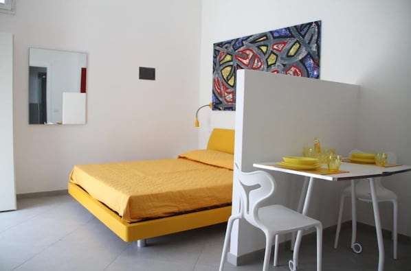A bedroom with a yellow bed, white chairs, a small table with yellow items, and a colorful abstract painting on the wall.