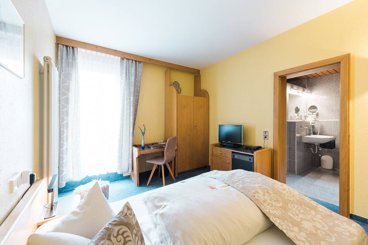 Quarto de hotel com cama, escrivaninha com computador, televisão, banheiro com pia e espelho, e janela com cortinas.