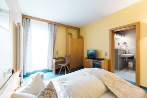 Una habitación de hotel con una cama, un escritorio con computadora, televisor, baño con lavamanos y espejo, y una ventana con cortinas.