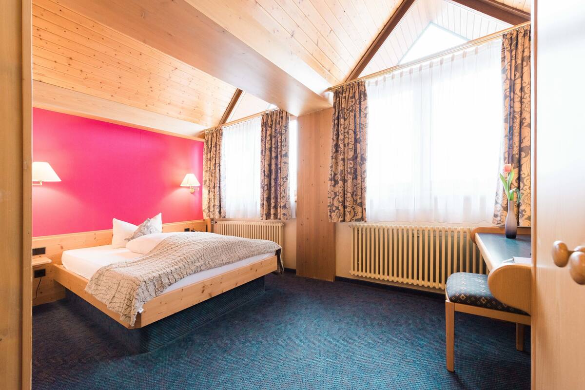 Quarto com cama, escrivaninha e cadeira. Duas janelas com cortinas, um radiador e um abajur na parede.