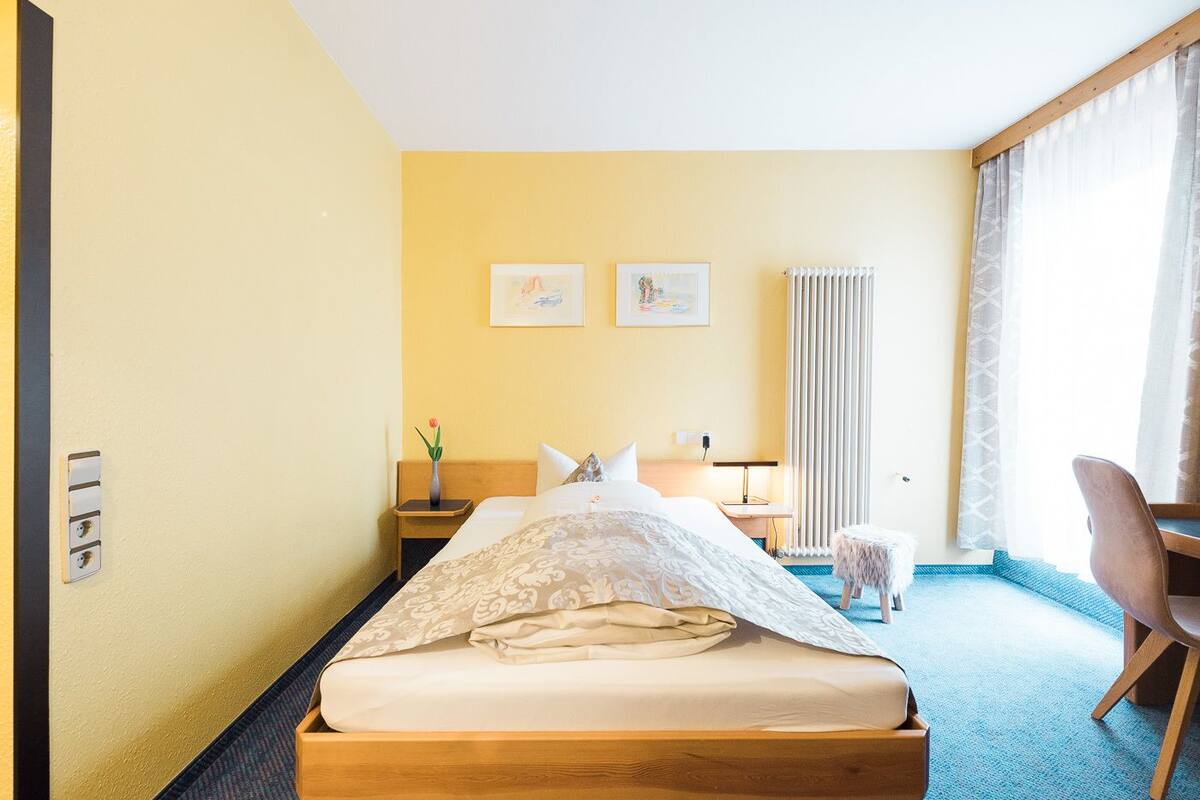 Quarto de hotel com cama, escrivaninha, cadeira e janela com cortinas.