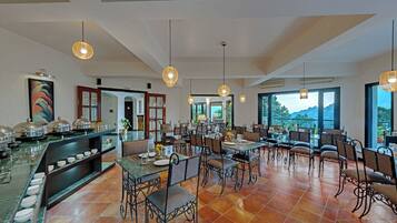 Un amplio comedor con mesas y sillas altas, un mostrador de buffet con platos, y grandes ventanales que ofrecen vistas al exterior.
