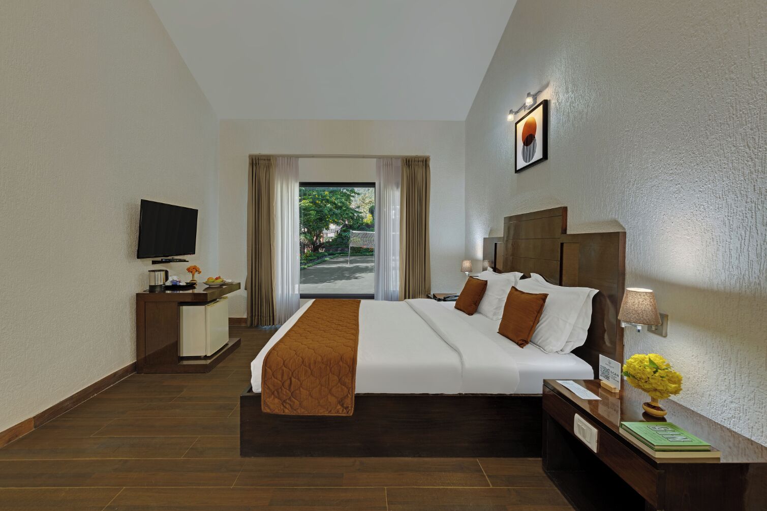 Habitación de hotel con una cama grande, un escritorio con televisor de pantalla plana, una mesita de noche con lámpara y un jarrón de flores, y vista al verdor exterior.