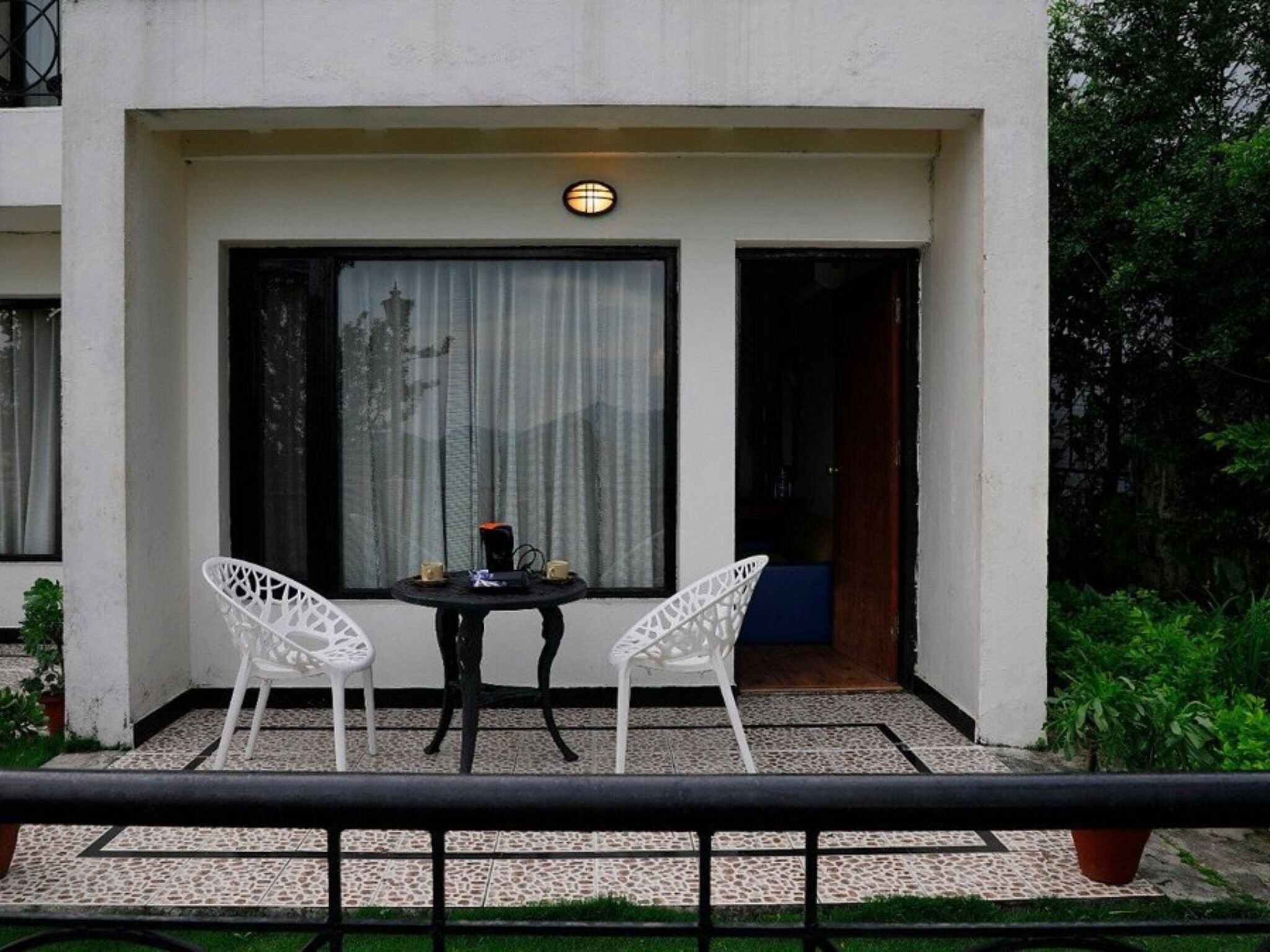 A small outdoor seating area with two white chairs and a table, a window with curtains, and a door leading inside.