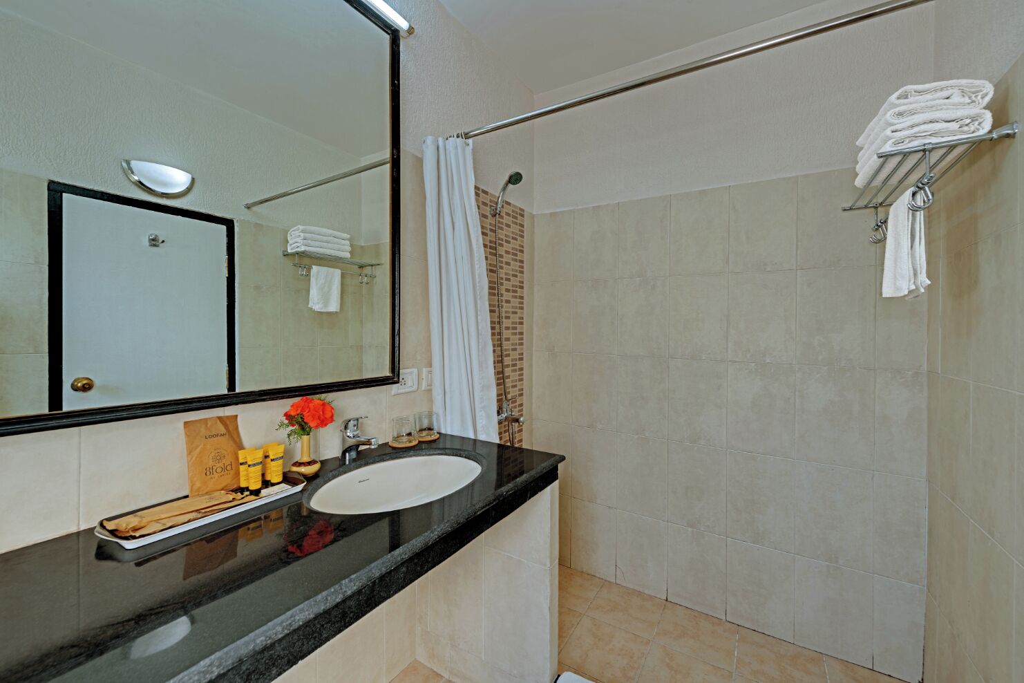 A bathroom with a black countertop, white sink, and a large mirror. There is a shower curtain, a towel rack, and a small shelf with toiletries.