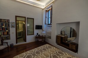 Una stanza con pavimento in legno, una libreria, una scrivania con lampada, uno specchio e una finestra con tende.
