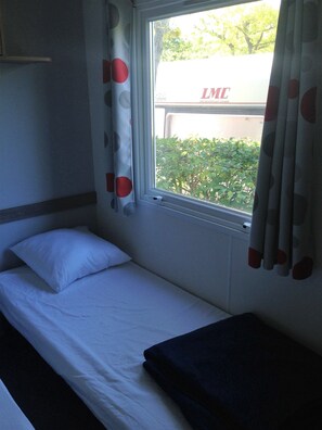 Una cama individual con ropa blanca y una almohada oscura, una ventana con cortinas y vistas al exterior con vegetación.