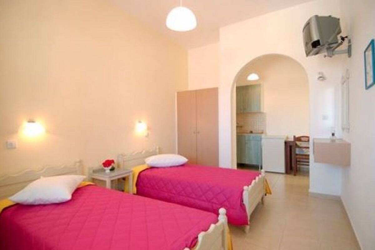 A room with two beds, a small table with a vase, and a kitchenette in the background.