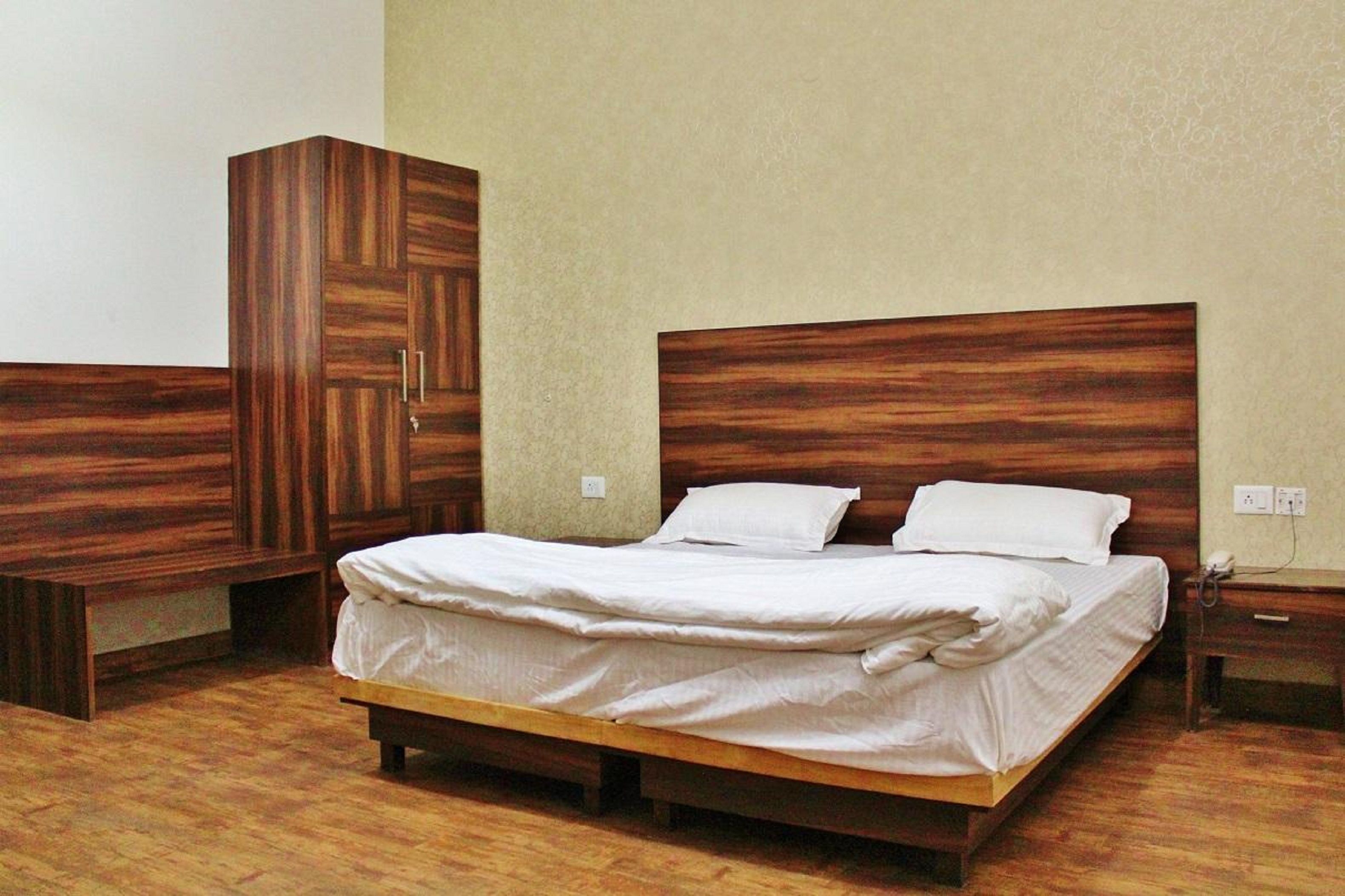 Een slaapkamer met een houten bedframe, twee witte kussens, een houten garderobe en een nachtkastje met een telefoon.