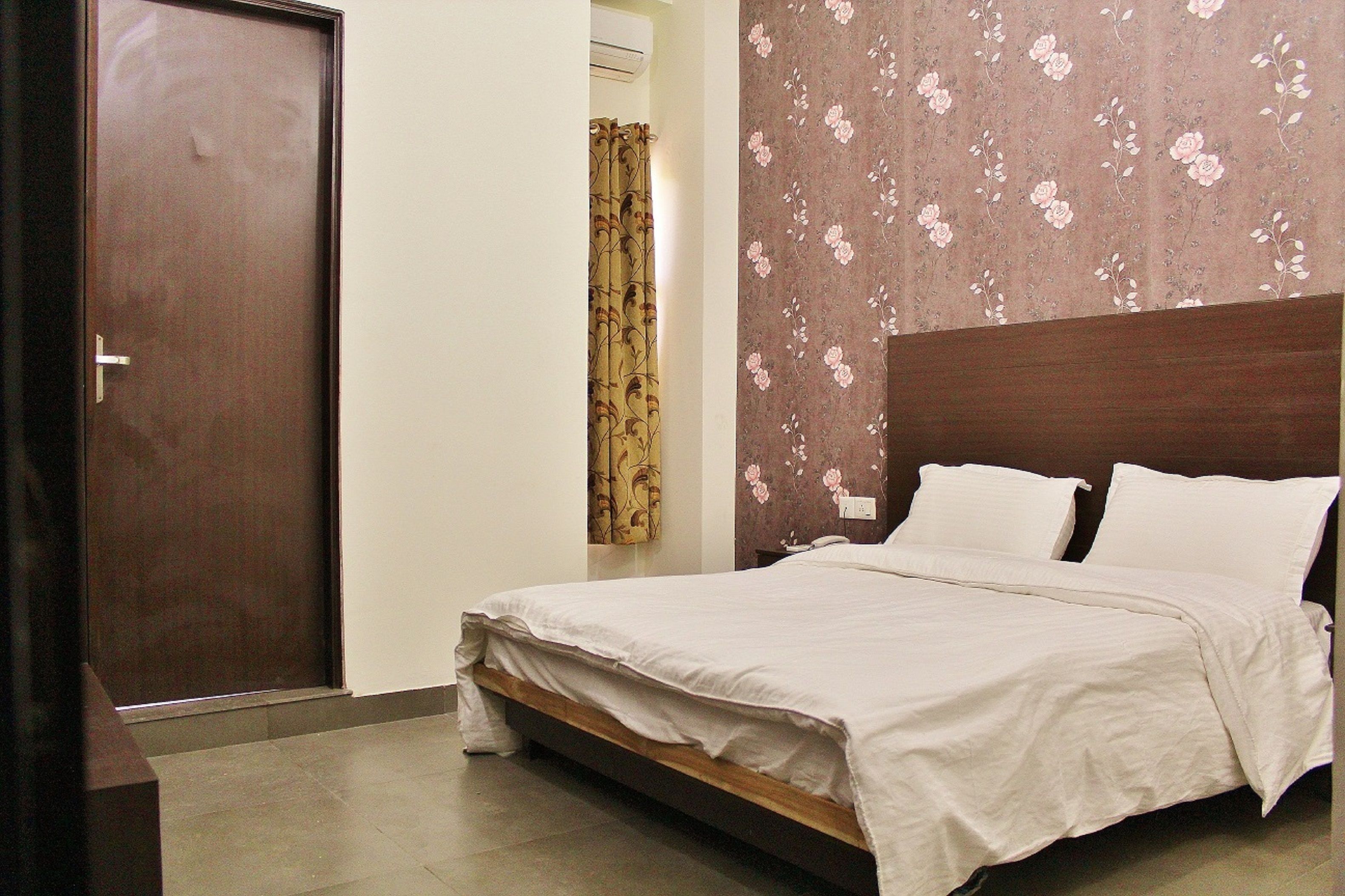 Un dormitorio con estructura de cama de madera, ropa de cama blanca, pared con estampado floral y una puerta cerrada.
