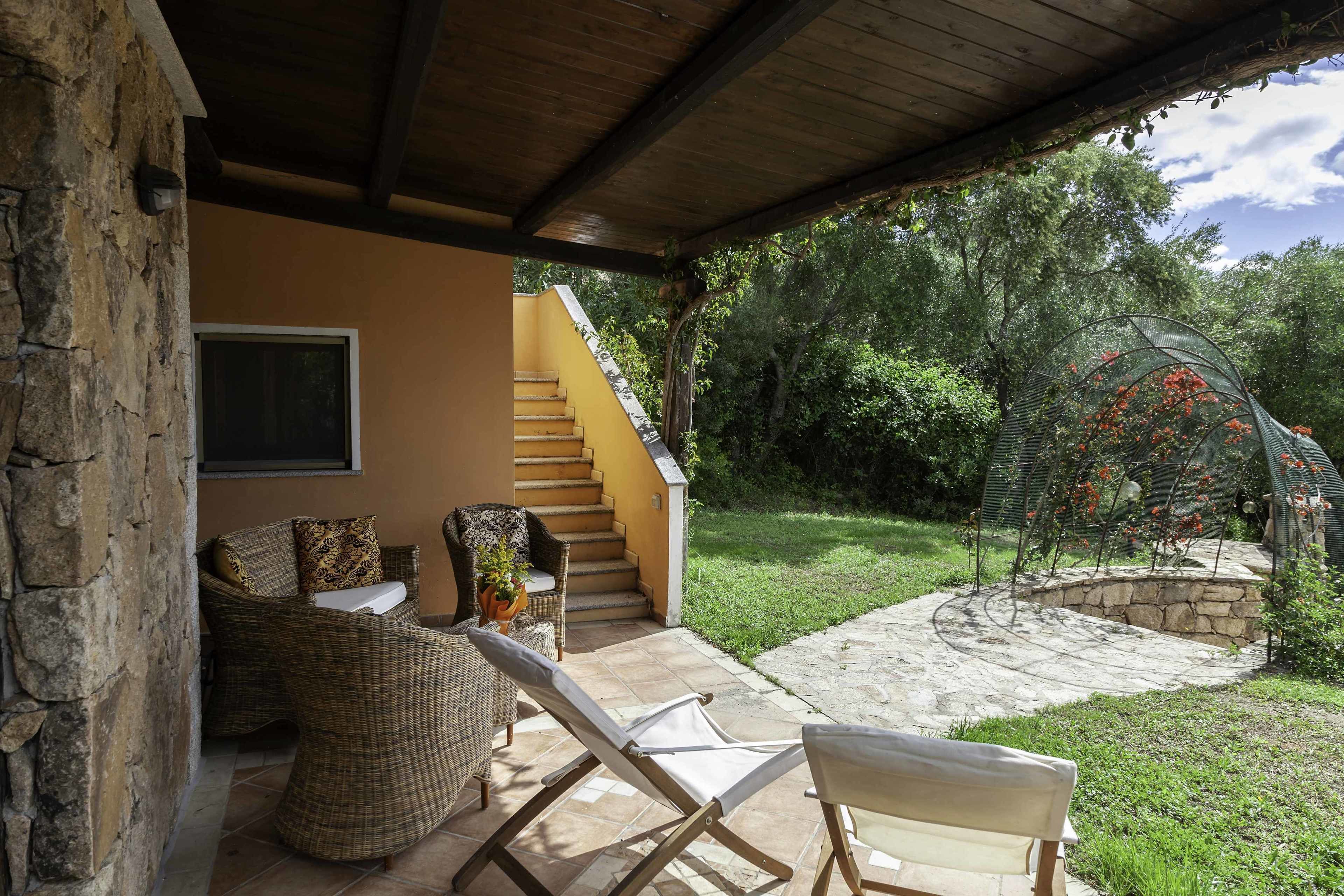 A covered outdoor seating area with wicker furniture, a stone wall, and a staircase leading to an upper level.