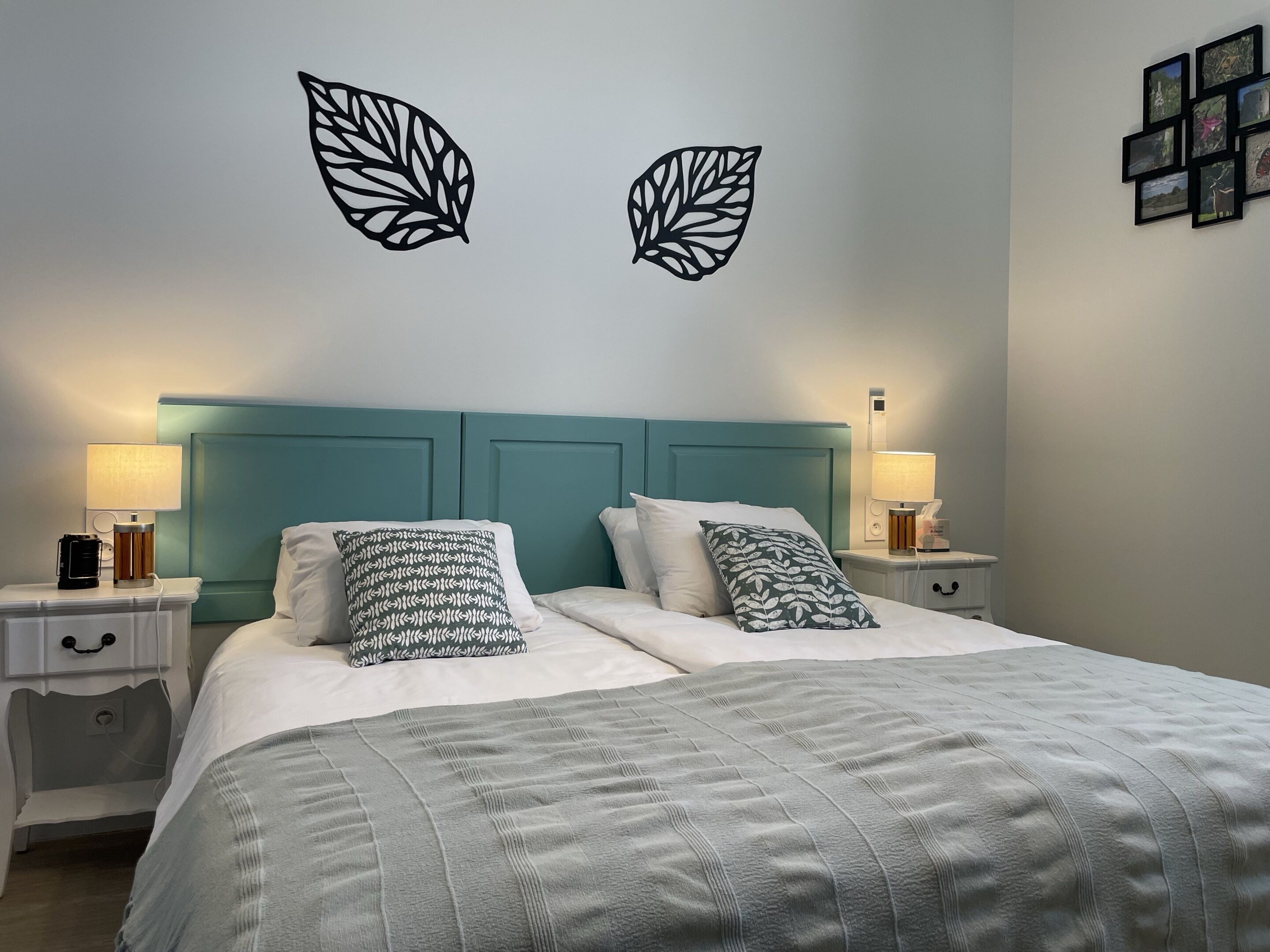 A neatly made double bed with a teal headboard, two bedside tables with lamps, and decorative leaf wall art.