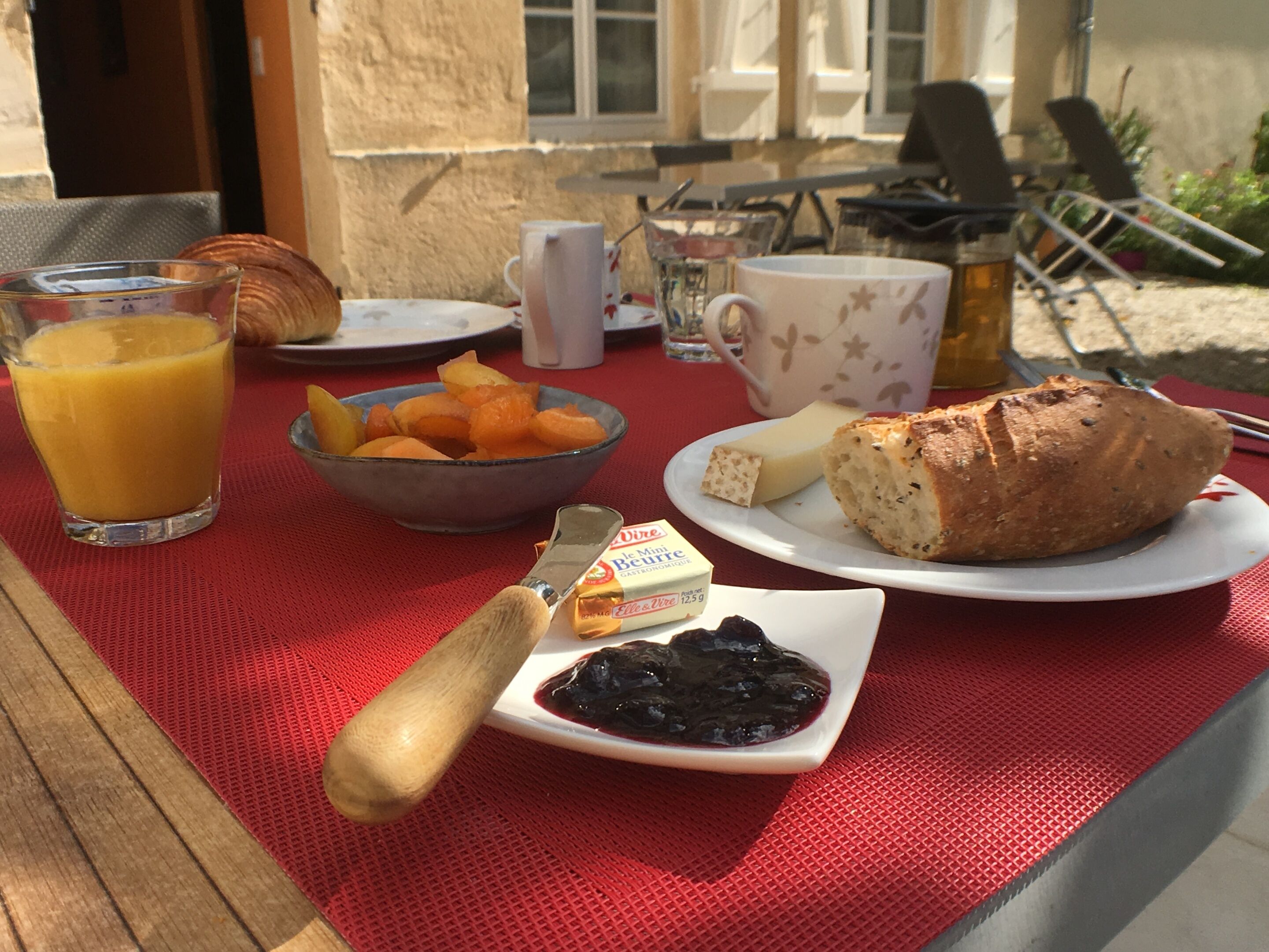 A breakfast table with a glass of orange juice, a bowl of fruit, a croissant, cheese, and a baguette.
