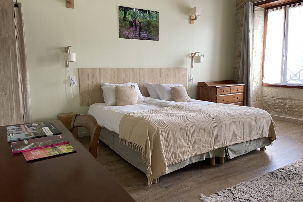 Un dormitorio con cama de madera, mesitas de noche, una mesita auxiliar, una ventana con cortinas y un cuadro colgado en la pared.