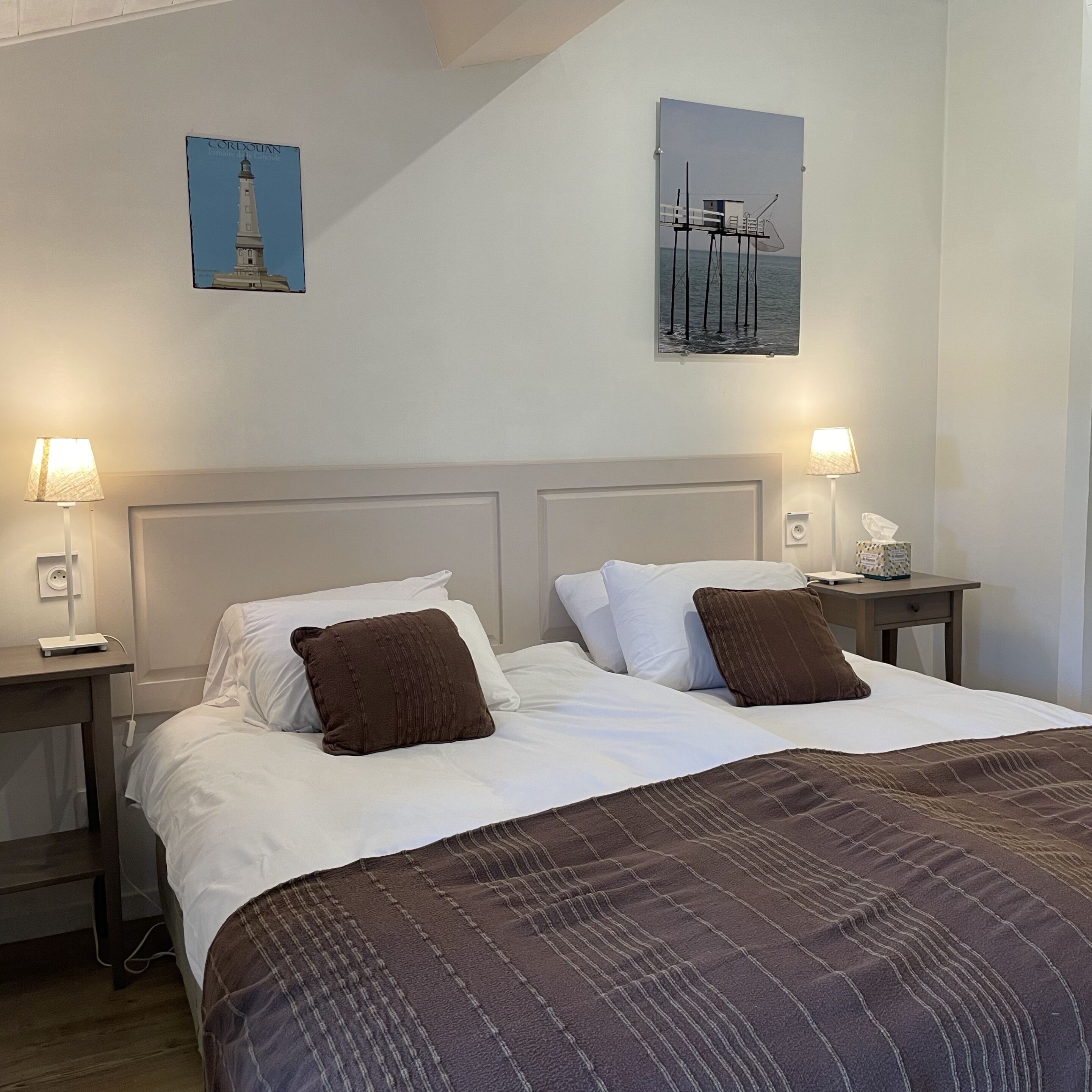 Habitación de hotel con dos camas, cada una con sábanas blancas y almohadas marrones. Hay dos mesitas de noche con lámparas y un teléfono. Las paredes son lisas y decoradas con dos cuadros enmarcados.
