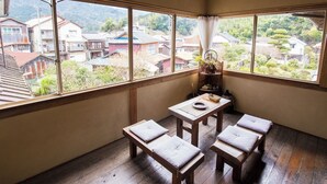 A room with a wooden table and benches, a view of houses through large windows, and a traditional Japanese shelf with items.